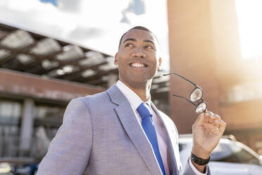 Happy businessman holding eyeglasses on sunny day - JCCMF05670