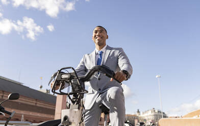 Glücklicher Geschäftsmann auf einem Elektrofahrrad sitzend an einem sonnigen Tag - JCCMF05673