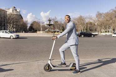 Smiling businessman standing with electric push scooter on sunny day - JCCMF05678