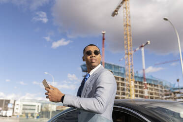 Geschäftsmann mit Sonnenbrille und Smartphone am Auto stehend an einem sonnigen Tag - JCCMF05683