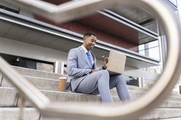 Lächelnder Geschäftsmann mit Laptop auf den Stufen vor einem Gebäude sitzend - JCCMF05699