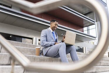 Smiling businessman using laptop sitting on steps in front of building - JCCMF05699