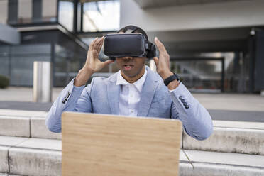 Businessman wearing virtual reality simulator sitting with laptop on steps - JCCMF05702