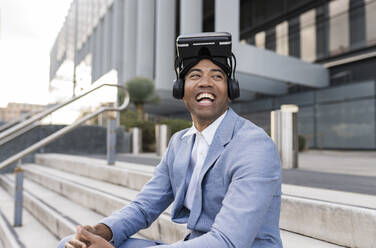 Cheerful businessman wearing virtual reality simulator sitting in front of building - JCCMF05707