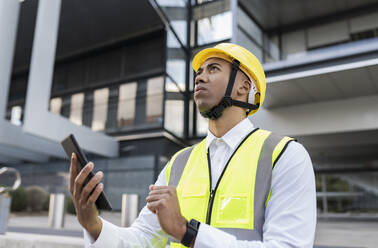 Man with hardhat holding tablet PC standing in front of building - JCCMF05708