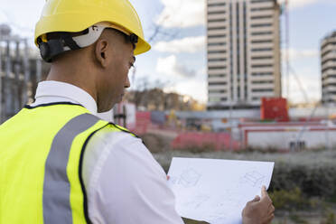 Architect wearing hardhat reading blueprint at construction site - JCCMF05714