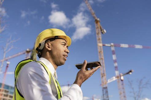 Ingenieur, der auf einer Baustelle an einem sonnigen Tag über einen Lautsprecher mit seinem Smartphone spricht - JCCMF05719