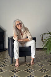 Woman wearing eyeglasses sitting on chair in front of white wall - VEGF05411