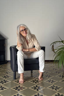 Frau mit Brille sitzt auf einem Stuhl vor einer weißen Wand - VEGF05411