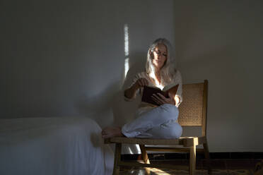 Eine Frau liest ein Buch auf einem Stuhl am Bett zu Hause - VEGF05414