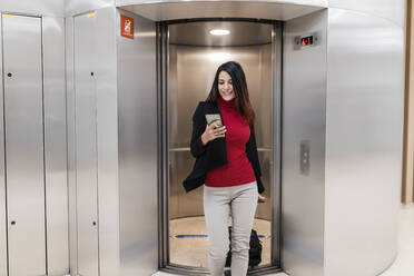 Businesswoman using smart phone moving out from elevator at office corridor - JRVF02872
