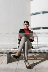 Lächelnde Geschäftsfrau mit Laptop auf einer Bank sitzend - JRVF02884