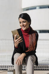 Smiling businesswoman using smart phone sitting on bench - JRVF02887