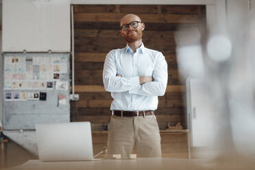 Smiling businessman with arms crossed standing in office - JOSEF07592