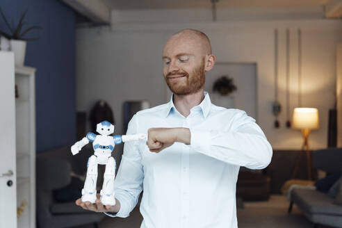 Smiling businessman gesturing fist bump with toy robot standing in office - JOSEF07604