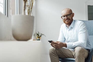 Smiling businessman with mobile phone sitting on chair in office - JOSEF07610