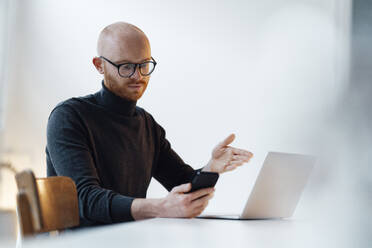 Young businessman using smartphone sitting with laptop working in office - JOSEF07615