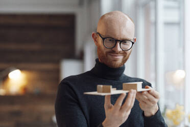 Smiling young businessman with wooden model in office - JOSEF07640