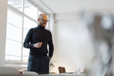 Smiling young businessman standing with hand in pocket at desk - JOSEF07643