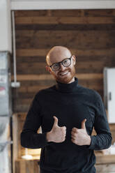 Happy young working man gesturing thumbs up in office - JOSEF07650