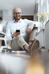 Lächelnder junger Geschäftsmann, der ein Mobiltelefon benutzt und auf einem Skateboard im Büro sitzt - JOSEF07653