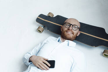 Lächelnder Geschäftsmann mit Mobiltelefon auf dem Skateboard im Büro liegend - JOSEF07656