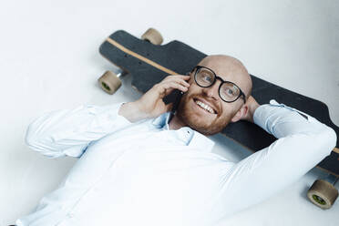 Glücklicher Geschäftsmann, der mit einem Mobiltelefon spricht und auf einem Skateboard im Büro liegt - JOSEF07658