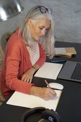 Smiling freelancer with gray hair writing on notepad at home - VEGF05417