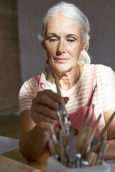 Woman holding painting brush sitting at table - VEGF05433