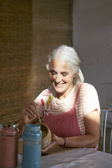 Smiling artist painting clay pot sitting on chair - VEGF05434