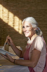 Lächelnde Frau, die am Tisch sitzend eine Tonplatte bemalt - VEGF05437