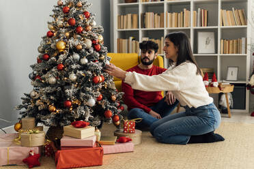 Junger Mann sitzt bei seiner Freundin und schmückt den Weihnachtsbaum zu Hause - GIOF14905