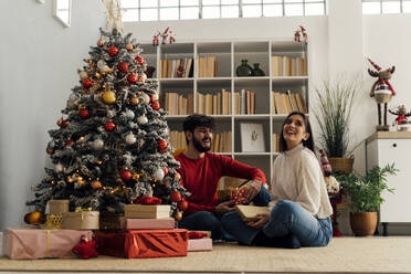 Glückliche Frau mit Freund, der eine Geschenkbox hält, sitzt zu Hause am Weihnachtsbaum - GIOF14906