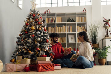 Glückliches Paar, das eine Geschenkbox hält, die unter dem Weihnachtsbaum zu Hause steht - GIOF14907