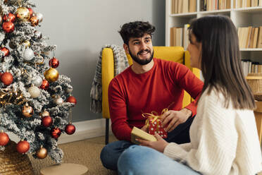 Glücklicher Mann mit Freundin, die eine Geschenkbox hält, sitzt zu Hause unter dem Weihnachtsbaum - GIOF14909