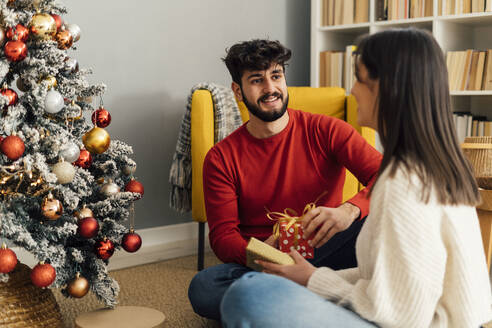 Glücklicher Mann mit Freundin, die eine Geschenkbox hält, sitzt zu Hause unter dem Weihnachtsbaum - GIOF14909
