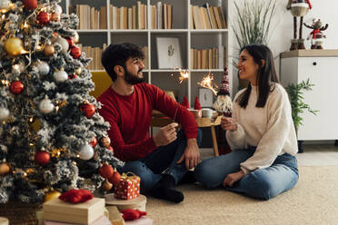 Smiling couple holding lit sparklers in living room at home - GIOF14912