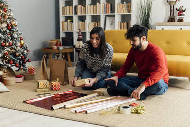 Happy couple sitting with Christmas presents and wrapping paper at home - GIOF14921