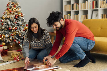 Smiling couple wrapping Christmas present in living room at home - GIOF14922