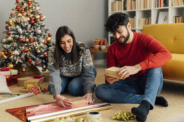 Happy couple wrapping Christmas present sitting in front of sofa - GIOF14923