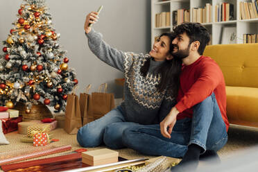 Lächelndes Paar, das ein Selfie mit seinem Smartphone am Weihnachtsbaum zu Hause macht - GIOF14924