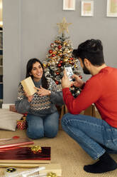 Man photographing girlfriend holding gift box sitting in front of Christmas tree - GIOF14925