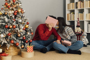 Mann versteckt Gesicht mit Geschenk-Box und küssen Freundin sitzt im Wohnzimmer - GIOF14927