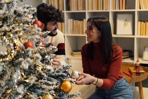 Lächelnde junge Frau hält Weihnachtsschmuck und sieht ihren Freund an - GIOF14943