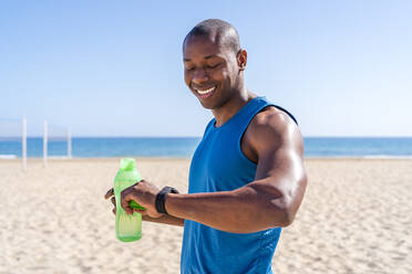 Lächelnder Mann mit Wasserflasche, der die Zeit am Strand an einem sonnigen Tag überprüft - DLTSF02803