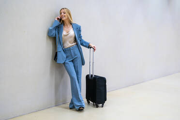 Lächelnde Geschäftsfrau, die mit einem Smartphone spricht und mit Gepäck auf Rädern vor einer Wand steht - DLTSF02820