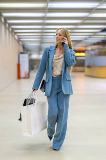 Lächelnde blonde berufstätige Frau, die mit dem Handy spricht und mit Einkaufstaschen in der U-Bahn-Station spazieren geht - DLTSF02821