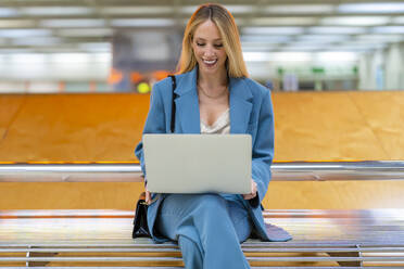 Glückliche arbeitende Frau mit Laptop auf einer Bank in einer U-Bahn-Station sitzend - DLTSF02825