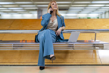Lächelnde blonde Geschäftsfrau, die mit ihrem Laptop auf einer Bank in einer U-Bahn-Station sitzt und mit einem Handy spricht - DLTSF02826