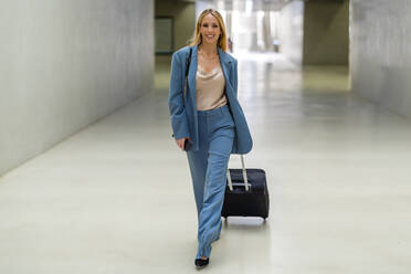 Happy blond businesswoman with wheeled luggage walking at subway station - DLTSF02828